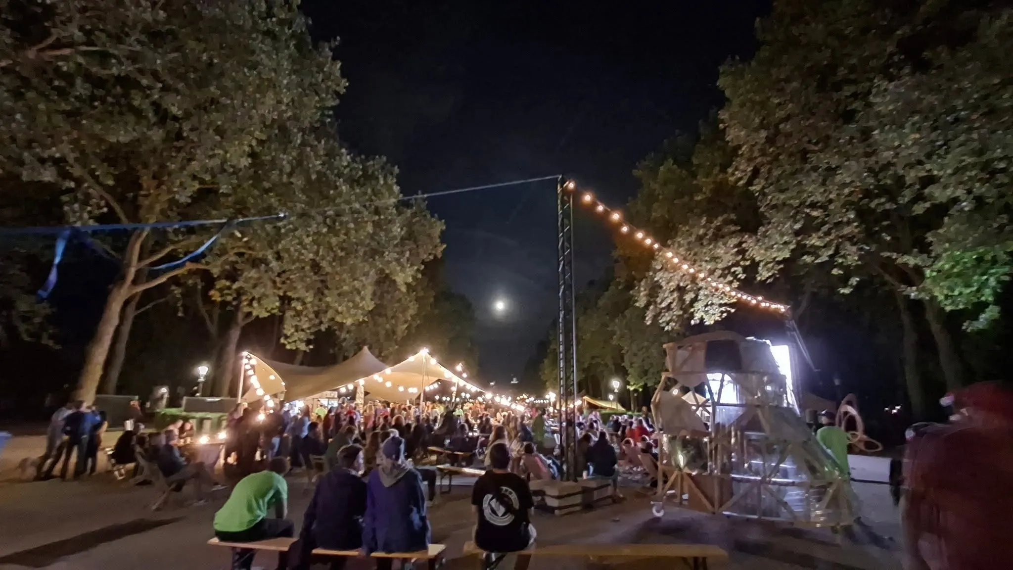 Het festivalterrein bij nacht. Slingers met lampjes tussen de bomen, mensen aan de picknicktafels, de maan boven het park.