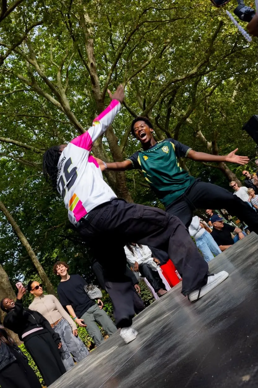 Twee breakdancers battelen op een podium tussen de bomen, het publiek juicht mee.