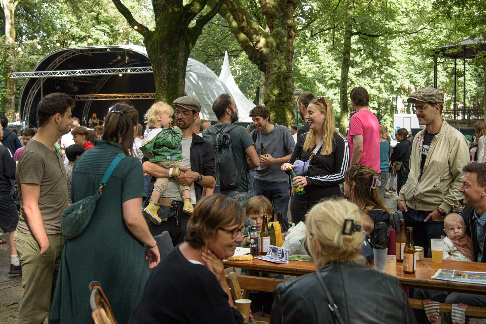 Des festivaliers aux tables de pique-nique près de la scène, familles et guirlandes dans les arbres.